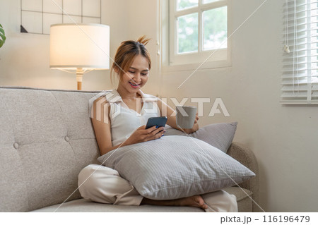 Young Woman Relaxing on Sofa with Smartphone and Coffee in Cozy Living Room Young Woman Relaxing on Sofa with Smartphone and Coffee in Cozy Living Room 116196479
