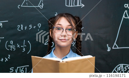 Beautiful teenage schoolgirl reading a textbook for a teacher while standing at a blackboard at school 116198171