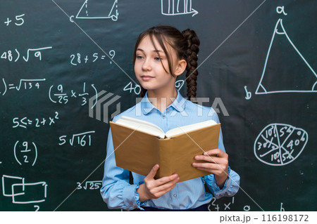 Beautiful teenage schoolgirl reading a textbook for a teacher while standing at a blackboard at school 116198172