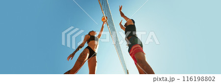 Two people playing beach volleyball, reaching for ball above t net, against clear blue sky. Banner. Active and dynamic hobby 116198802