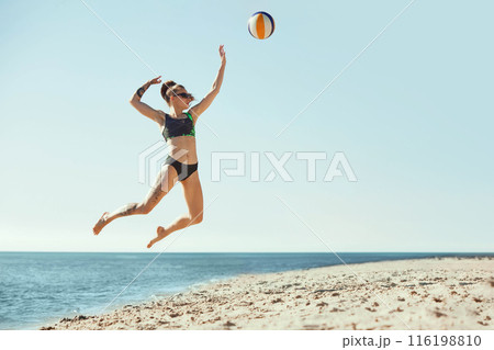 Full-length dynamic image of young woman in sunglasses and s swimwear playing beach volleyball, hitting ball in a jump. Blue sky and sea background. Full-length dynamic image of young woman in sunglasses and s swimwear playing beach volleyball, hitting ball in a jump. Blue sky and sea background. 116198810