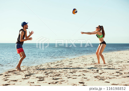Two players in action on sunny beach, playing volleyball under clear blue sky by tranquil sea. Dynamic man and woman during game 116198852