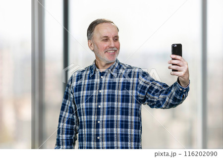Portrait of smiling mature man taking a selfie on his smartphone camera. Standing indoors on windows backgorund. Portrait of smiling mature man taking a selfie on his smartphone camera. Standing indoors on windows backgorund. 116202090