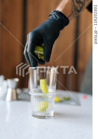 Vertical cropped shot of unrecognizable professional bartender in black gloves putting kiwi slices into glass to make deliciously refreshing cocktail in summer cafe. Concept of celebration mood. Vertical cropped shot of unrecognizable professional bartender in black gloves putting kiwi slices into glass to make deliciously refreshing cocktail in summer cafe. Concept of celebration mood. 116202801
