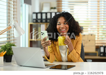 Cheerful female entrepreneur relaxing at workplace, using mobile phone and drinking coffee Cheerful female entrepreneur relaxing at workplace, using mobile phone and drinking coffee 116202978
