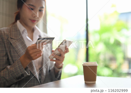 Young businesswoman holding a credit card making online payment on mobile phone, sitting at workplace table 116202999