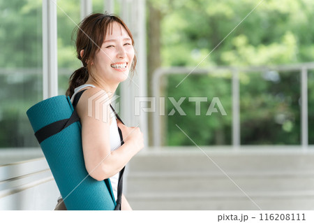 ヨガマットを持って笑顔で振り返るヨガウェア姿の若いアジア人女性 116208111