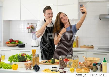 Young couple lover cooking together in the kitchen at home. Happy couple taking selfie with smartphone or making video call. 116213418