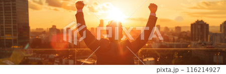 A person celebrating success on a rooftop at sunrise with a cityscape in the background, arms raised in victory. 116214927
