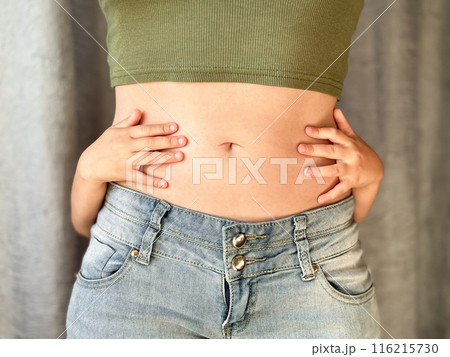 Close up of woman in jeans and green top with childs hands around her waist. Concept of family bonding, motherhood, postpartum recovery and affection. Neutral background 116215730