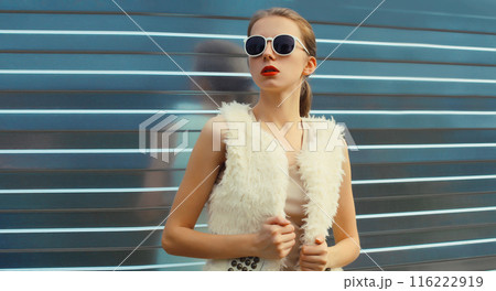 Modern stylish young woman in white fur vest, glasses, fashionable girl posing on gray background Modern stylish young woman in white fur vest, glasses, fashionable girl posing on gray background 116222919