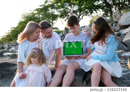 A family of five sits with a laptop in the mountains near the ocean Cheerful family with kids on beautiful summer meadow learning on laptop Family laptop green screen 116223348
