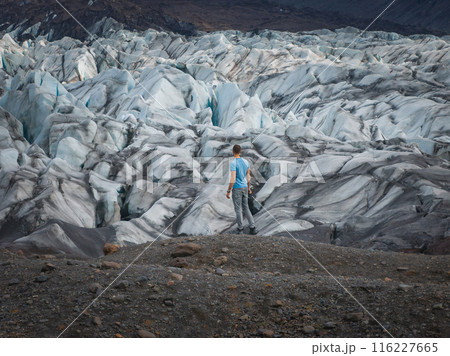 A man stands on a rocky outcropping, dwarfed by a vast glacier in Iceland. The glacier's crevasses and ice formations stretch out as far as the eye can see. 116227665