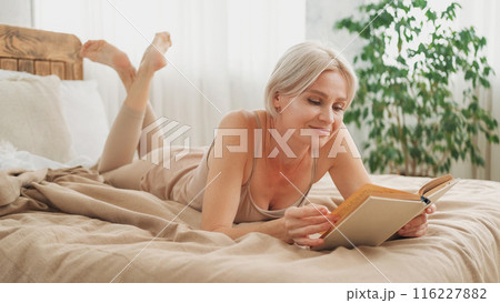 Enjoying reading. Home rest. Happy peaceful relaxed woman in sleeping suit laying with novel book on comfortable bed in light hygge interior. 116227882