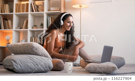 Video chat. Virtual communication. Relaxed cheerful girl in headphones using laptop enjoying online conversation on floor at light home interior with free space. 116227904