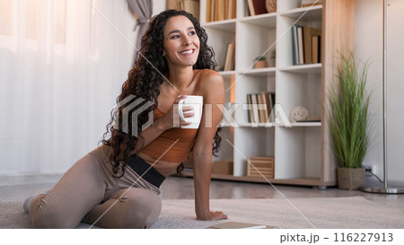Morning coffee. Home leisure. Weekend rest. Happy relaxed smiling woman with mug daydreaming enjoying hot drink on floor at modern living room interior with free space. 116227913