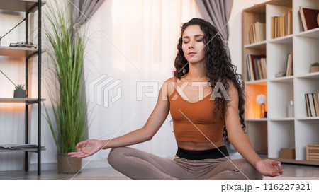 Home yoga. Indoor meditation. Health wellbeing. Relaxed tranquil woman sitting cross-legged in lotus pose at light modern living room interior with free space. 116227921
