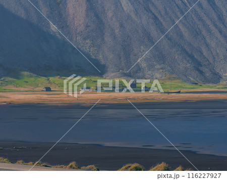 A wide shot capturing the contrast between a dark, rocky mountainside and a small, green village on a flat, grassy area in Iceland. Unique landscape. 116227927