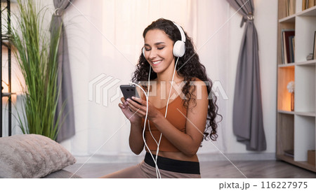 Music app. Phone playlist. Gadget entertainment. Relaxed smiling woman in headphones enjoying listening to song at light modern home interior with free space. 116227975