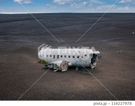 A white, partially destroyed airplane with bullet holes sits on a black sand beach in Iceland. The beach is empty and the sky is blue with white clouds. 116227978