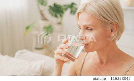 Water balance. Good morning. Healthy habit. Happy woman drinking from glass enjoying hydration in light living room interior at home copy space. 116228043