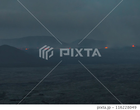 A breathtaking aerial shot capturing a dark, misty volcanic landscape in Iceland, with glowing red lava flows visible in the distance. 116228049