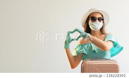 Pandemic love. Heart gesture. Girl in gloves mask summer dress sunglasses strew hat with suitcase showing hand affection sign isolated on white empty space background. 116228301