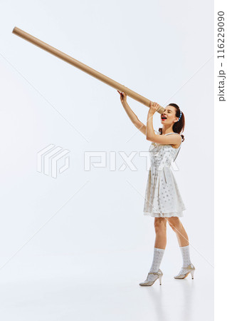Young woman in polka dot dress and vintage shoes looking into giant cardboard tube isolated on white background. Looking for innovation and inspiration. Young woman in polka dot dress and vintage shoes looking into giant cardboard tube isolated on white background. Looking for innovation and inspiration. 116229090