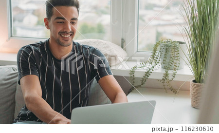 Online communication. Internet chatting. Happy smiling casual man typing on laptop enjoying virtual connection sitting on couch in light living room interior at home copy space. 116230610