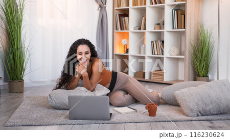 Online leisure. Internet fun. Relaxed amused cheerful girl enjoying watching movie on laptop on floor at modern home living room interior with free space. 116230624