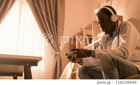Doctor break. Digital leisure. Mobile entertainment. Male physician in headphones enjoying playing video game online on phone at modern home interior with copy space. Doctor break. Digital leisure. Mobile entertainment. Male physician in headphones enjoying playing video game online on phone at modern home interior with copy space. 116230699