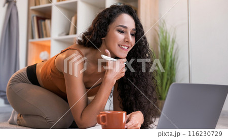Internet research. Online learning. Distance education. Curious cheerful girl watching video lesson using laptop taking notes at modern home interior with copy space. Internet research. Online learning. Distance education. Curious cheerful girl watching video lesson using laptop taking notes at modern home interior with copy space. 116230729