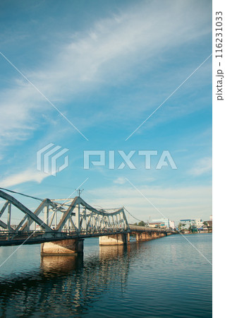 A calm river flows under a the famous old bridge in Kampot town in Cambodia, reflecting the clear blue sky and scattered clouds, with a cityscape in the background A calm river flows under a the famous old bridge in Kampot town in Cambodia, reflecting the clear blue sky and scattered clouds, with a cityscape in the background 116231033