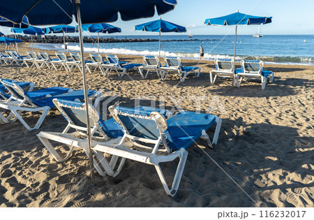 Beach sunbeds with blue umbrellas, chairs on sandy beach on Tenerife seaside resort, coastal town leisure 116232017