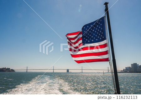 American flag on pole flutters against blue sky, Golden Gate Bridge visible in background over San Francisco Bay. City skyline faintly visible to left. Prideful composition. 116232113
