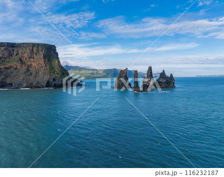 Stunning aerial image of Iceland's coastline featuring a large cliff and tall rock formations. Rising from the deep blue ocean under a bright blue sky. Stunning aerial image of Iceland's coastline featuring a large cliff and tall rock formations. Rising from the deep blue ocean under a bright blue sky. 116232187