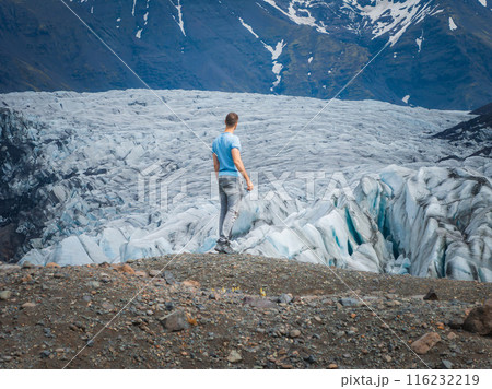 A man in a blue shirt stands on a rocky outcropping, gazing at a stunning glacier with icy blue and white hues in Iceland. The scene captures the awe-inspiring beauty and vastness of the Icelandic 116232219