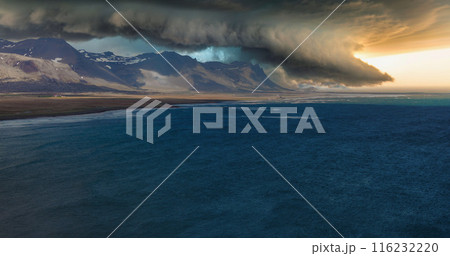 A dark storm cloud looms over a rugged, snow-covered mountain range in Iceland. The calm, dark blue ocean contrasts with the barren, desolate landscape. A dark storm cloud looms over a rugged, snow-covered mountain range in Iceland. The calm, dark blue ocean contrasts with the barren, desolate landscape. 116232220