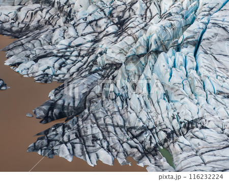 Stunning aerial image of an Icelandic glacier, showcasing intricate details of snow and ice with deep cracks, surrounded by brown, muddy water. 116232224