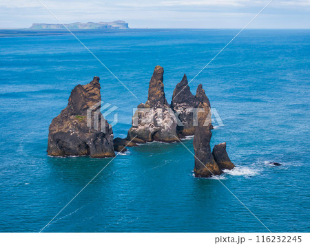 Dramatic aerial view of Reynisdrangar sea stacks off Iceland's southern coast, with black sand beach and distant coastline in the background. Dramatic aerial view of Reynisdrangar sea stacks off Iceland's southern coast, with black sand beach and distant coastline in the background. 116232245