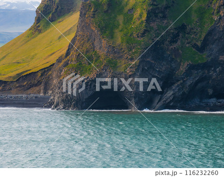 A stunning aerial view of Iceland's black sand beach featuring a large cave in a cliff face made of basalt columns. Deep blue water and light blue sky complete the scene. A stunning aerial view of Iceland's black sand beach featuring a large cave in a cliff face made of basalt columns. Deep blue water and light blue sky complete the scene. 116232260