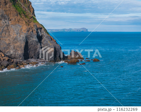 Aerial view of a rocky cliff covered in green vegetation and brown rock, jutting into the clear blue ocean with small rocks and islands in the distance. Aerial view of a rocky cliff covered in green vegetation and brown rock, jutting into the clear blue ocean with small rocks and islands in the distance. 116232269