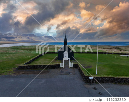 Aerial view of the black church in Budir, Iceland. The small wooden church with a white steeple is surrounded by a stone wall, grassy field, water, and mountains. 116232280