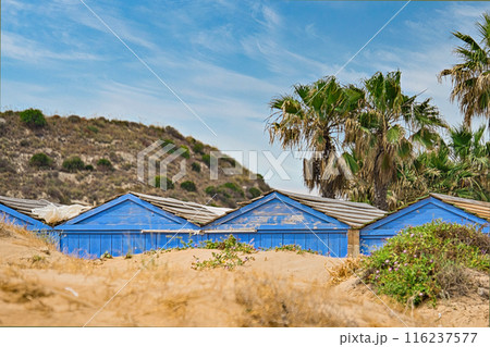 Weathered blue beach huts on sandy beach, drowning in dunes near lush palm trees and scenic hillsides under clear blue sky 116237577