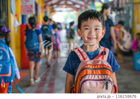 A young boy with a bright smile and a colorful backpack stands in a school hallway, surrounded by other students on their way to class. 116238676