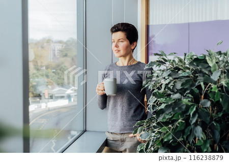 Middle aged minded pensive employee business woman relaxing with coffee cup and day dreaming while looking through the window from her office. Dreaming, planning, Taking time for recovery. Copy space 116238879