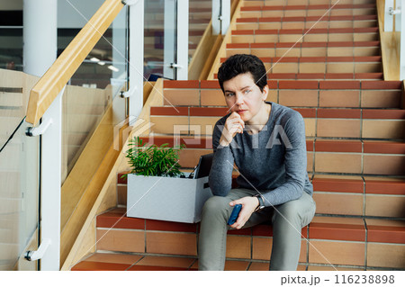 Stressed worried middle aged gender neutral woman with box of personal items sitting alone on the staircase after being fired, lost job due to recession and economic stress. Equality discrimination. 116238898