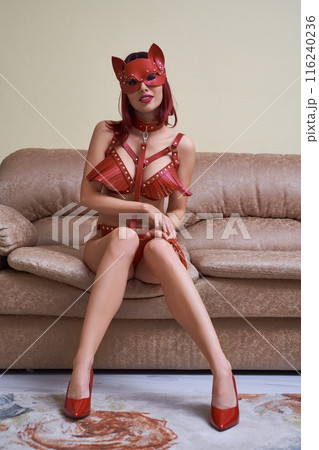 Sexy woman in red leather bondage and cat mask sitting on sofa 116240236