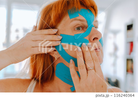 Close-up portrait middle-aged woman, she puts kinesiology tape on face Close-up portrait middle-aged woman, she puts kinesiology tape on face 116240364