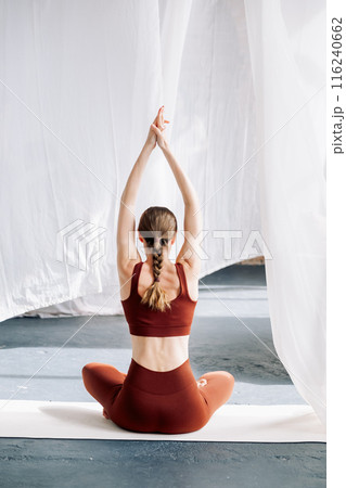 A slender woman in a brown sports uniform with a pigtail sits in an asana against the background of white curtains and meditates, rear view. Yoga classes in the loft studio A slender woman in a brown sports uniform with a pigtail sits in an asana against the background of white curtains and meditates, rear view. Yoga classes in the loft studio 116240662